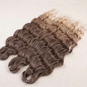May include: A set of three hair extensions featuring a wavy texture and an ombre colour gradient. The hair transitions from a dark brown shade at the top to a lighter, golden blonde tone at the ends, with each bundle secured by a small metal clip.