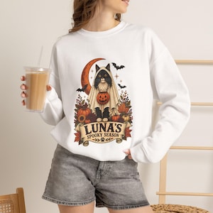 Può includere: Una felpa bianca a maniche lunghe con la grafica di un gatto nero vestito da fantasma, che tiene in mano un secchiello a forma di zucca. Il design include una luna crescente, pipistrelli, foglie autunnali e la scritta LUNA'S SPOOKY SEASON.