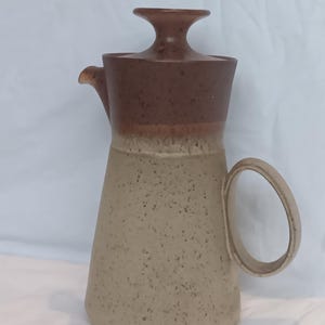 Könnte beinhalten: Ein Keramikkrug mit zweifarbigem Design, bestehend aus einem dunkelbraun gesprenkelten oberen Teil und einem helleren, beige gesprenkelten Unterkörper. Der Krug verfügt über einen passenden Deckel mit zentralem Knauf, einen kleinen Ausguss und einen runden Henkel.