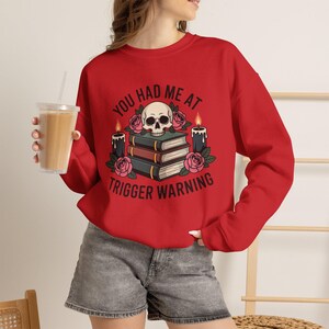 Puede incluir: Una sudadera roja de manga larga con un gráfico de una calavera sobre una pila de tres libros, rodeada de rosas rosas y dos velas negras. El texto "YOU HAD ME AT TRIGGER WARNING" está impreso en negro alrededor de la ilustración central.