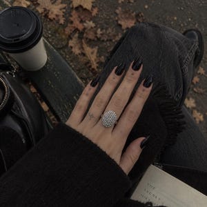 May include: A silver-toned ring featuring a large, oval-shaped face densely encrusted with numerous small, clear, sparkling stones. The ring is worn on a finger, showcasing a prominent, textured, and reflective surface that catches the light.