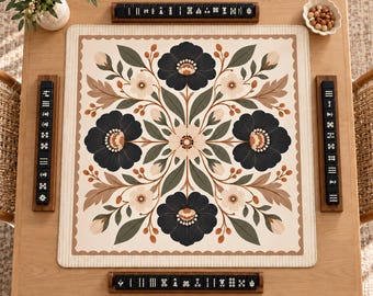 May include: A square mahjong mat featuring a symmetrical floral pattern in dark blue, sage green, and cream, surrounded by four wooden tile racks holding black mahjong tiles with white symbols, arranged on a light wood table.