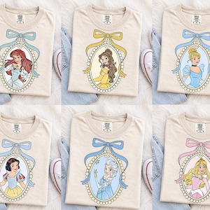 Puede incluir: Una colección de seis camisetas beige, cada una con una ilustración diferente de una princesa de dibujos animados en un marco ovalado de cuentas rematado con un lazo de cinta de color. Los personajes incluyen a Ariel, Bella, Cenicienta, Blancanieves, Elsa y Aurora.