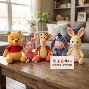 Peut inclure: Un ensemble de cinq animaux en peluche crochetés comprenant un ours jaune en chemise rouge, un petit cochon rose, un tigre orange à rayures noires, un âne bleu et un lapin jaune. Un panneau blanc affiche des drapeaux et le texte Available Languages.