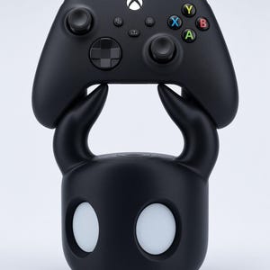 Könnte beinhalten: Ein mattschwarzer Gaming-Controller-Ständer in Form einer stilisierten Figur mit zwei geschwungenen Hörnern und zwei kreisförmigen, weißen augenähnlichen Aussparungen. Ein schwarzer kabelloser Controller mit weißem Logo liegt auf den Hörnern.