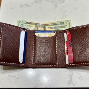 May include: A dark brown leather trifold wallet with visible white stitching, featuring three interior card slots and a central compartment holding a twenty-dollar bill. The centre card slot is embossed with a circular logo and text.