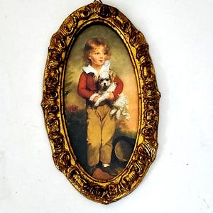 May include: A framed oval portrait of a young boy holding a small white dog. The frame is gold with ornate details. The portrait is set against a white background.