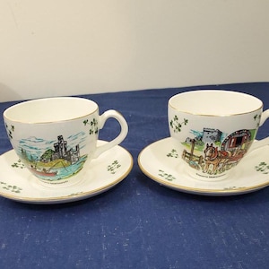 May include: Two white teacups with gold trim and shamrock designs. One cup features a scene of a castle and a boat, the other a horse-drawn cart. Both cups are on matching saucers with shamrock designs.
