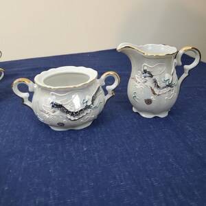 May include: A white porcelain sugar bowl and creamer set. The set features a dragon design with gold trim. The sugar bowl has two handles and a lid. The creamer has a handle and a spout. The dragon design is in black, blue, and pink.