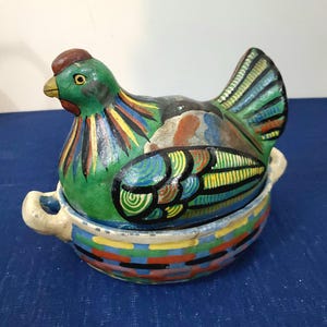 May include: A hand-painted ceramic hen-shaped container. The hen is primarily green with colorful accents on the wings and body. The base is a bowl with a striped pattern in various colors. The container has two small handles.