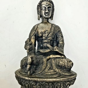 May include: A silver-colored metal statue of Buddha sitting in a meditative pose. The statue is detailed with intricate carvings and is sitting on a base with a decorative design.