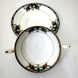 May include: White porcelain soup bowl and saucer set. The bowl has two handles and a gold rim. The rim and saucer feature a decorative border with a black background, green and orange floral design.