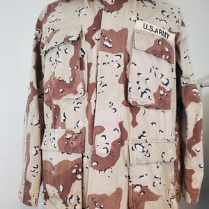May include: A tan, brown, and black camouflage military jacket with a U.S. Army patch on the right chest.