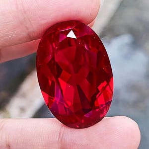 May include: A large, oval-cut gemstone with a deep, vibrant red color and a faceted surface that reflects light. The stone displays a high level of clarity and a brilliant, polished finish, showcasing a symmetrical shape and intense saturation.