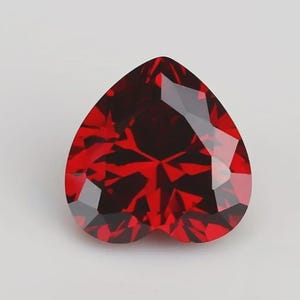 Puede incluir: Una piedra preciosa en forma de corazón de color rojo intenso y vibrante con una superficie facetada. La piedra presenta múltiples facetas triangulares y en forma de diamante que crean un patrón complejo de luz y sombra.