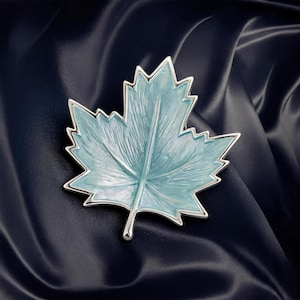 May include: A maple leaf-shaped dish featuring a glossy light blue enamel interior with visible textured vein details and a polished silver-toned metal border that outlines the entire shape of the leaf, including the stem at the bottom.