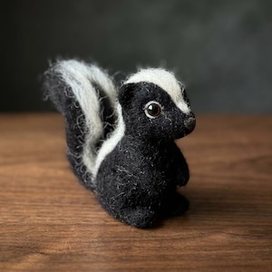 Peut inclure: Une petite figurine de mouffette en laine feutrée à l'aiguille, composée de fibres noires et blanches. La figurine présente un corps noir texturé, une bande blanche proéminente du sommet de la tête au dos, une queue touffue et deux yeux sombres.