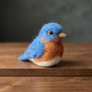 May include: A needle-felted bird figurine featuring a bright blue head and back, a warm orange-brown chest, and a white belly. The bird has a small, pointed brown beak and a single visible dark, glossy eye, crafted with a soft, textured wool finish.