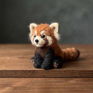 May include: A needle-felted red panda figurine featuring a textured rust-orange and brown coat, a cream-coloured face with black accents around the eyes, a small black nose, and dark black limbs. The small animal is depicted in a seated position.