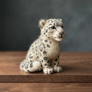 以下が含まれることがあります： ニードルフェルトで作られたユキヒョウの子供のフィギュア。クリーム色の毛並みに濃いグレーと黒の斑点模様が施されています。丸みを帯びた頭部、尖った耳、羊毛の質感、そして暗く光沢のある目を持つ、座った姿勢の小さな彫刻作品です。