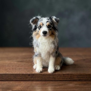 Puede incluir: Una figura de perro Pastor Australiano hecha con lana fieltrada con aguja, que presenta un pelaje azul mirlo con parches de pelo blanco, negro y canela. La escultura tiene una apariencia texturizada y lanosa, ojos oscuros y nariz negra.
