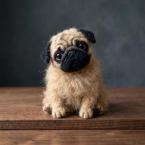 May include: A needle-felted pug figurine with a textured tan body and a dark black muzzle and ears. The small dog sculpture features large, glossy black eyes and a soft, fuzzy appearance, capturing the distinct facial features of a pug.