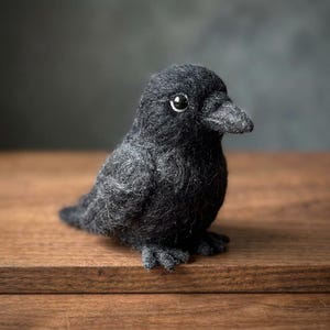May include: A needle-felted black bird figurine with a textured, fuzzy wool surface. The bird has a small, pointed beak, a rounded head, and a single visible dark eye with a white highlight, capturing a realistic avian form in a compact size.