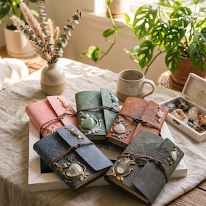 May include: Five leather-bound journals in pink, green, brown, blue, and dark gray, each featuring embossed maple leaf patterns, a leather cord wrap, and a unique polished gemstone accent attached to the front cover with metal filigree.
