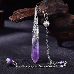 May include: A pointed, faceted purple amethyst crystal pendant encased in an ornate silver-toned metal cap. The pendant is attached to a silver-toned metal chain featuring a small round amethyst bead at the end and a textured silver sphere bead.