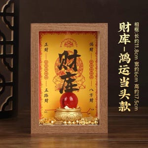 Op de afbeelding: Een rechthoekige houten lijst met een gele achtergrond met zwarte Chinese kalligrafie, een rode bol op een goudkleurige voet en kleine goudkleurige blokjes. De lijst meet ongeveer 11,8 cm bij 6 cm bij 17,5 cm.