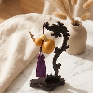May include: A dark brown wooden stand with a curved, dragon-like silhouette holds a hanging ornament. The ornament features a light yellow, double-gourd-shaped object, a small carved floral bead, and a vibrant purple tassel with a decorative knot.