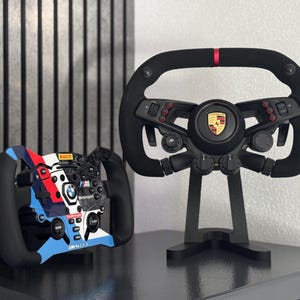 Könnte beinhalten: Ein schwarzes Rennlenkrad mit Porsche-Logo und roter Mittenmarkierung sowie ein kleineres Rennlenkrad mit blau-rot-weißer BMW Motorsport-Lackierung, ausgestattet mit verschiedenen Tasten, Drehreglern und integrierten Digitalanzeigen.