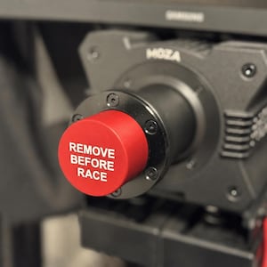 Könnte beinhalten: Eine schwarze Moza-Rennlenkradbasis mit einer auffälligen roten, runden Mittelkappe, auf der vorne der weiße Text REMOVE BEFORE RACE aufgedruckt ist. Das Gerät hat eine matte Oberfläche mit sichtbaren Schrauben und einem zylindrischen Anschluss.