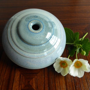 Può includere: Un vaso in ceramica blu chiaro con un'apertura rotonda e larga e un solo foro al centro. Il vaso ha una finitura testurizzata e smaltata e si trova su una superficie di legno scuro. Due fiori bianchi con centri gialli sono davanti al vaso.