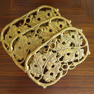 Könnte beinhalten: Set aus drei goldfarbenen Metall-Topfuntersetzern mit einem Blattdesign. Die Topfuntersetzer sind übereinander gestapelt.