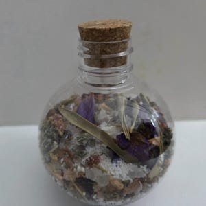 May include: A clear, spherical glass bottle with a cork stopper, filled with a mixture of white salt, dried purple flower petals, green leaves, and various small brown botanical elements, creating a textured, layered appearance inside the container.