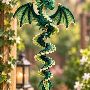 Könnte beinhalten: Ein hängendes Häkel-Drachenornament aus dunkelgrünem und hellgrünem Garn mit goldfarbenen Akzenten. Der Drache hat einen gewundenen Körper, detaillierte Flügel und ist an einer Kette mit einer facettierten grünen Kristallperle befestigt.