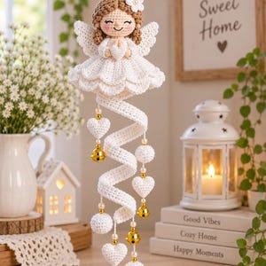 Puede incluir: Un adorno de ángel tejido a ganchillo con cabeza de hilo marrón claro, vestido blanco con bordes festoneados y halo dorado. Debajo del ángel, cuelga una cola en espiral de ganchillo blanco con corazones, esferas y cascabeles dorados.