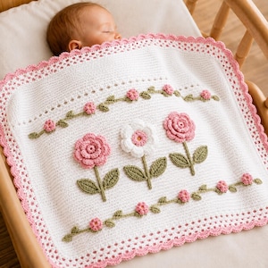 May include: A white crochet baby blanket featuring a scalloped pink border, two horizontal vine patterns with small pink flowers, and a central row of three large three-dimensional flowers in shades of pink and white with green leaves.