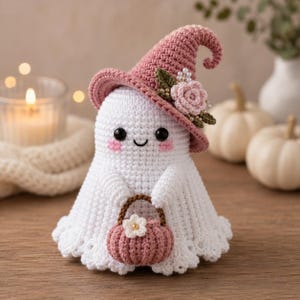 Pode incluir: Uma figura de fantasma em crochê branco usando um chapéu pontudo rosa empoeirado decorado com uma rosa em crochê, folhas verdes e pequenas contas brancas. O fantasma tem olhos pretos, bochechas rosadas e segura uma pequena abóbora de crochê.