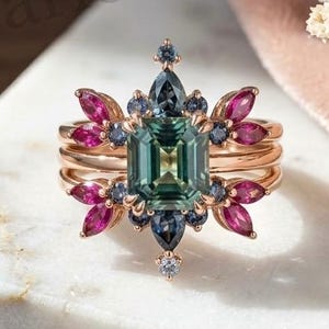 May include: A rose gold ring featuring a central emerald-cut green gemstone, surrounded by marquise-cut pink stones and round dark blue stones in a floral-inspired arrangement. The band is thin and polished, showcasing a symmetrical design.