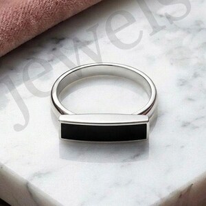 May include: A silver ring featuring a horizontal rectangular bar inlay of smooth black material. The ring has a polished metallic finish and a simple band design. The text a jewels is visible in the background behind the ring.