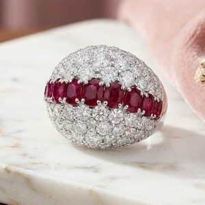 Op de afbeelding: Een koepelvormige ring met een centrale horizontale band van ovaal geslepen dieprode robijnen, geflankeerd door pavé gezette ronde witte diamanten. De ring is zilverkleurig. De tekst Farlea jewels is zichtbaar in het bovenste deel van de afbeelding.