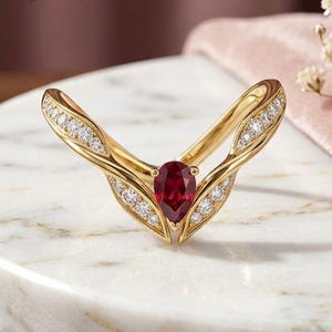 Op de afbeelding: Een gouden V-vormige ring met een peervormige rode robijn in het midden, geflankeerd door kleine ronde witte diamanten langs de gespleten ringband. De tekst Farlea jewels is zichtbaar in het bovenste gedeelte van de afbeelding.
