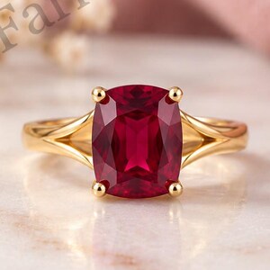 Op de afbeelding: Een gouden ring met een dieprode edelsteen in kussenslijpvorm, vastgehouden door vier pootjes. De ringband heeft een gespleten ontwerp dat aansluit op de zetting. De tekst Farlea jewels is zichtbaar in het bovenste deel van de afbeelding.