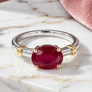 Puede incluir: Un anillo con un rubí rojo intenso de corte ovalado en el centro, montado sobre una banda de plata pulida. El engaste incluye pequeños detalles en oro amarillo a ambos lados de la piedra, la cual está asegurada por cuatro garras de oro.