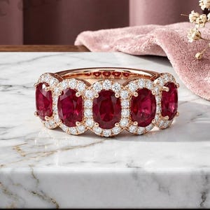 Peut inclure: Une bague en or rose ornée de cinq rubis rouge foncé taille ovale disposés en ligne. Chaque rubis est entouré d'un halo de petits diamants blancs ronds et clairs. L'anneau est fabriqué en or rose poli, présentant un design continu et élégant.
