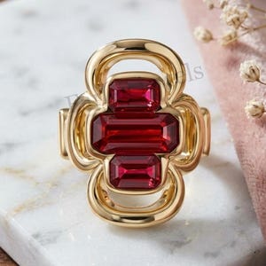 Peut inclure: Une bague dorée ornée de trois pierres rectangulaires rouge foncé à facettes, disposées verticalement. La monture en métal présente un design ajouré, lisse et arrondi, encadrant les pierres pour former une silhouette symétrique en trèfle.