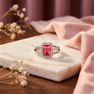 May include: A silver-toned ring featuring a large, rectangular, faceted red center stone. The central stone is surrounded by a halo of small, clear, round stones, with additional clear stones set along the split shank of the metal band.