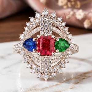 May include: A ring featuring a central radiant-cut red gemstone flanked by a pear-cut blue gemstone and a pear-cut green gemstone. The setting is surrounded by a halo of small white stones arranged in an oval, sunburst-like frame with pavé details.